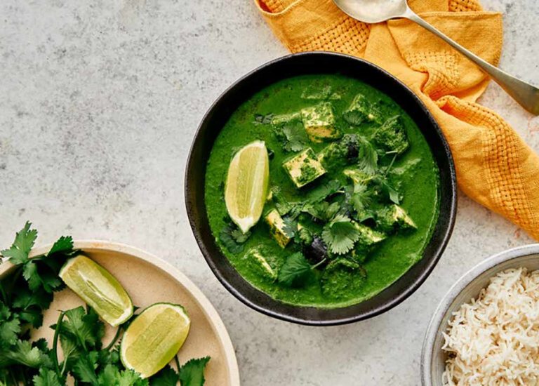 A bowl of green curry with plant-based paneer, coriander, and lime wedges—packed with iron-rich plants—sits next to a plate of limes, fresh coriander, a bowl of rice, and a yellow serviette with a spoon.