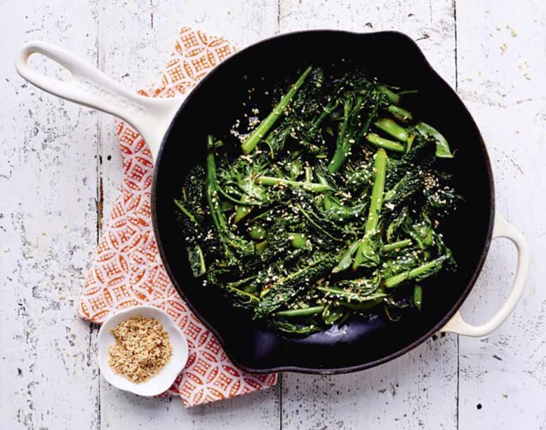A black frying pan with cooked broccolini—an iron-rich veg—topped with sesame seeds sits on a patterned tea towel. A small bowl of extra sesame seeds is nearby on a white timber surface, ideal for anyone wanting to boost their iron intake with tasty, plant-based choices.