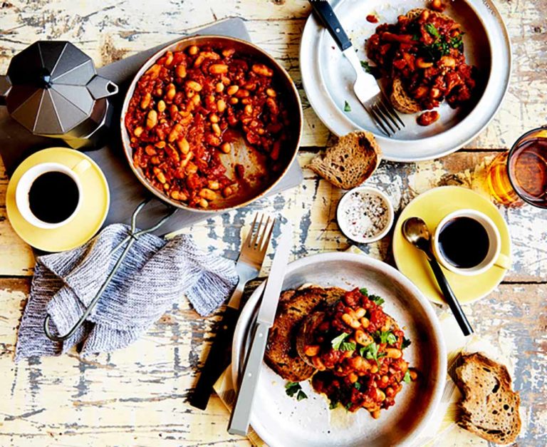 Two plates of toast topped with a hearty, plant-based iron bean stew sit beside a pot of the same rich stew, surrounded by coffee mugs, bread slices, cutlery, and a hob-top coffee maker on a rustic wooden table.