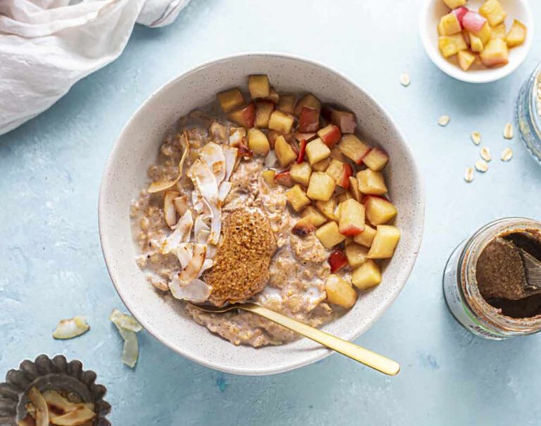 A bowl of porridge topped with diced apples, almond butter, and coconut flakes sits on a light blue surface with a gold spoon,