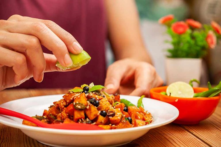 Hand squeezing a slice of lime over a plate of mixed veggie curry on a table, with a bowl of limes and green chillies and a small plant in the background.