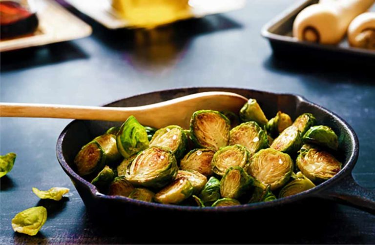 Sliced, roasted Brussels sprouts—one of the most iron-rich plant foods—are nestled in a black cast iron frying pan with a wooden spoon, sitting on a dark surface.