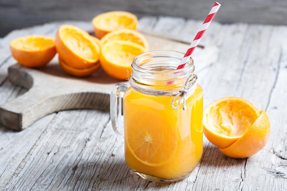 A jar of orange juice sits on a white wooden bench, with freshly juiced oranges scattered around. 