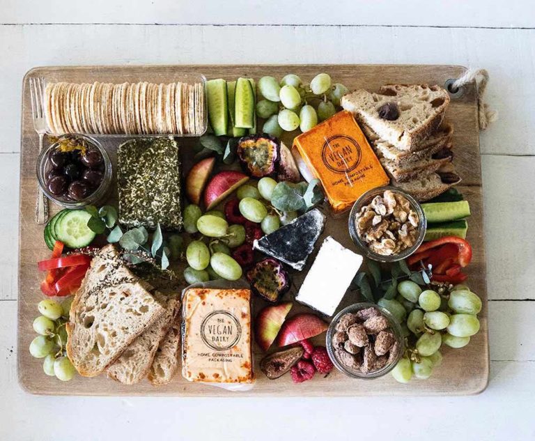 A wooden grazing board featuring a vegan cheese platter with crackers, sourdough bread, grapes, nuts, olives, and fresh vegetables.