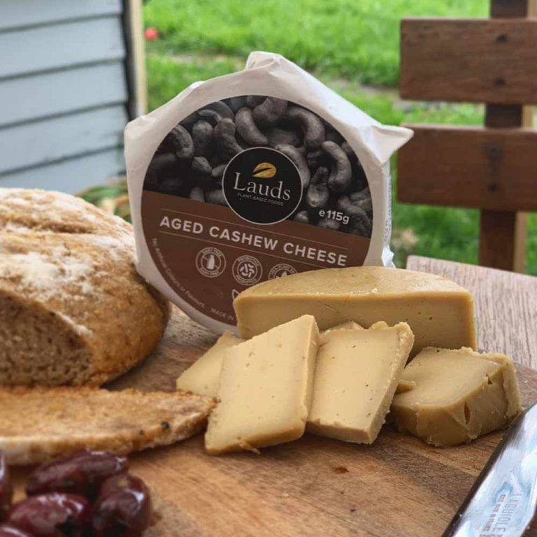 A round block of Lauds matured cashew cheese served on a plant-based cheese platter with crusty bread, olives, and several slices cut on a wooden board.