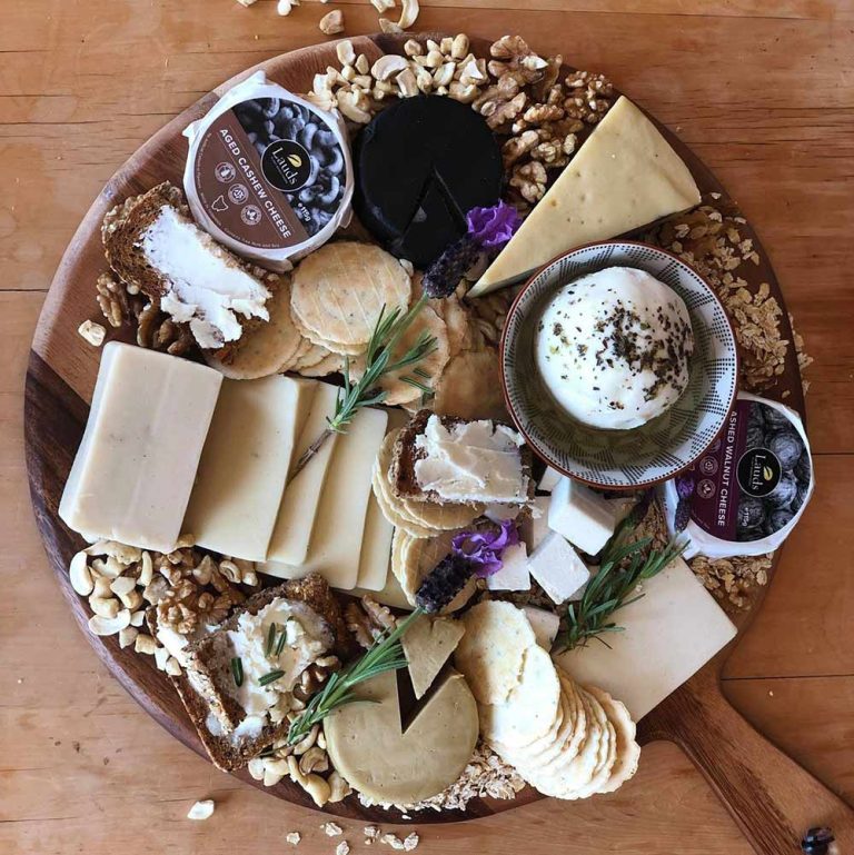 A wooden serving board featuring an assortment of plant-based cheeses, biscuits, walnuts, cashews, and herbs arranged on a vegan grazing platter.