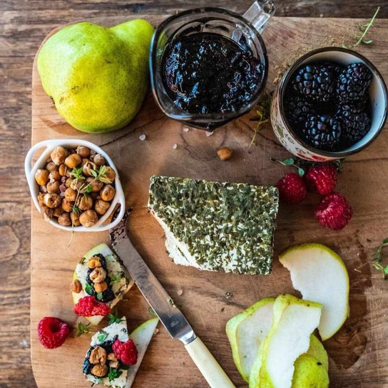 A wooden serving board featuring a plant-based cheese platter with herb cheese, pear slices, roasted chickpeas, blackberries, raspberries, and a jar of fruit preserve.