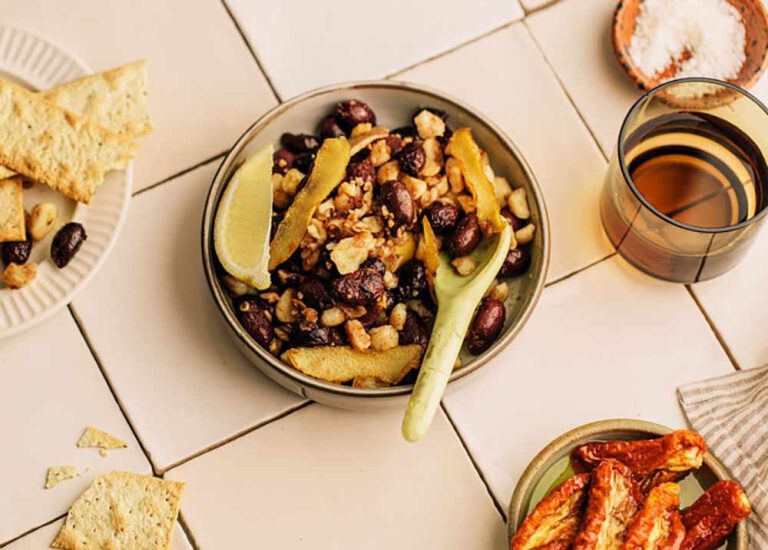 A bowl of olives, macadamias, and sun-dried tomatoes with lemon wedges and a spoon, served as a plant-based cheese platter with savoury biscuits.