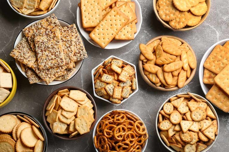 Multiple bowls filled with an assortment of plant-based crackers and pretzels for a dairy-free cheeseboard on a grey stone surface.
