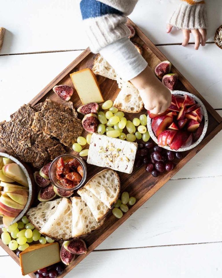 A child reaches for a piece of nectarine on a wooden serving board featuring a plant-based cheese platter, crackers, sourdough bread, grapes, figs, and sun-dried tomatoes.