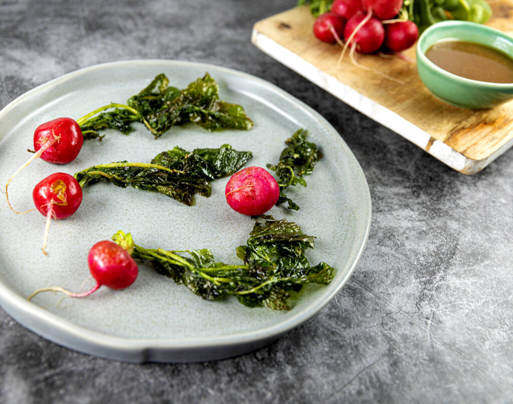 Three whole radishes and leaves on a grey plate, with more radishes and plant-based Apple Core Sauce on a wooden board behind.