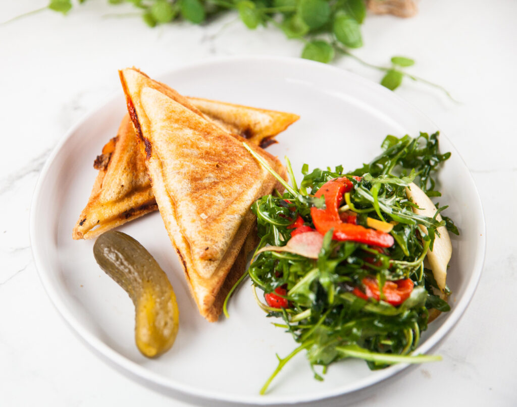 A white plate with two toasted plant-based Mushroom Ragu Jaffle halves, a pickle spear, and Rocket Salad with rocket and red peppers.