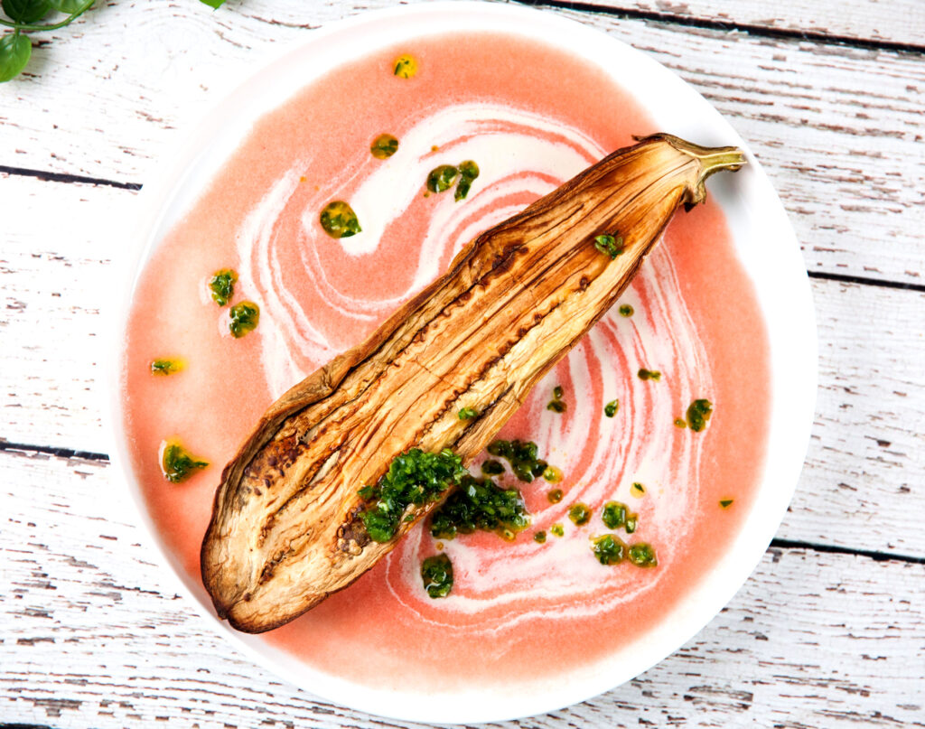 A halved, golden aubergine rests on pink and white plant-based creamy tahini sauce, topped with green herb oil on a white plate.