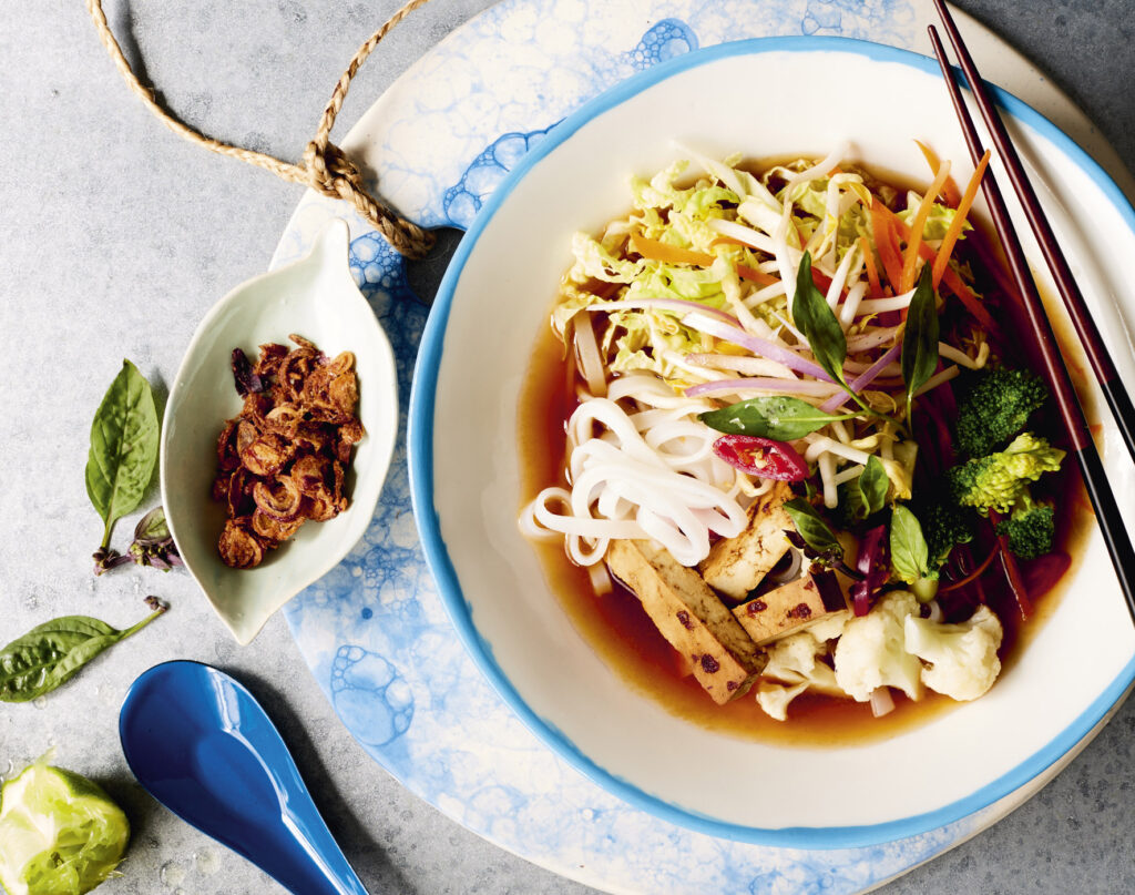 A bowl of plant-based pho with rice noodles, tofu, veg, ginger broth, fried shallots, chopsticks, and a blue soup spoon.