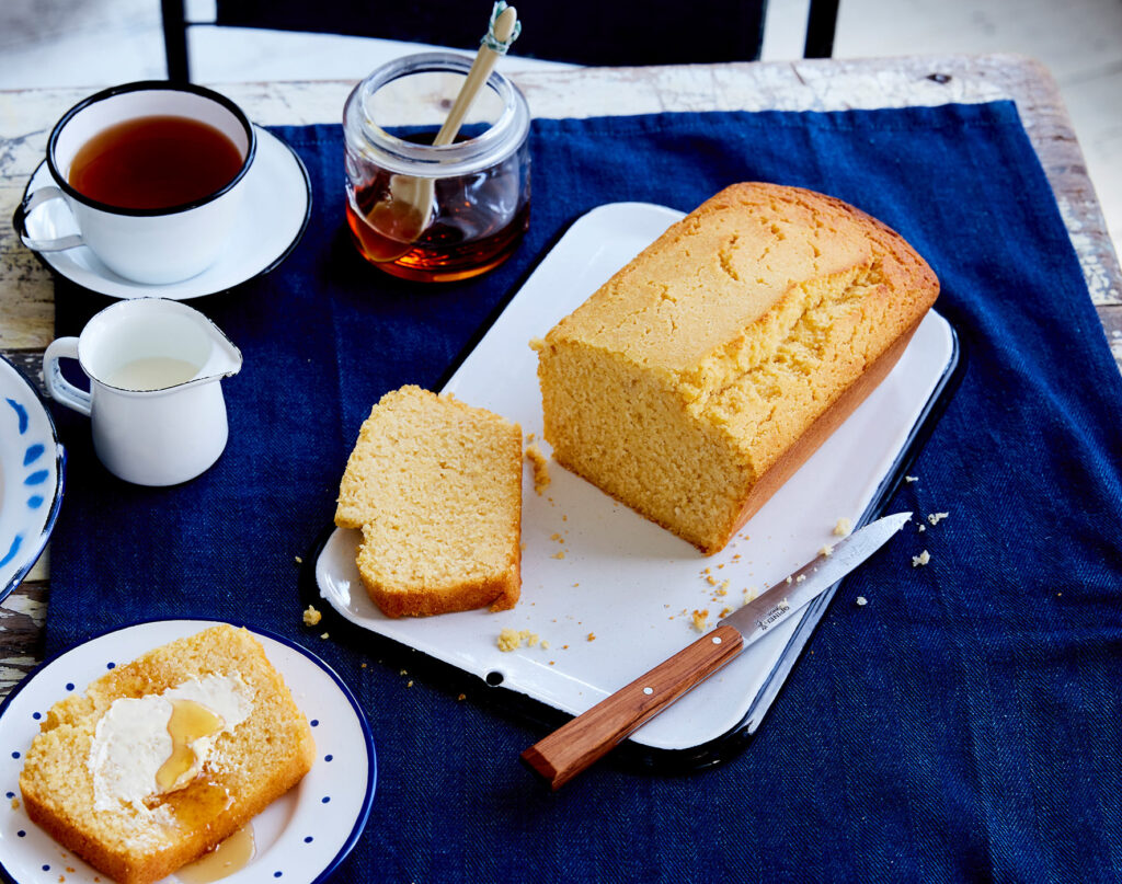 Golden cornbread is sliced on a tray with a knife, next to tea, plant-based honey, plant milk, and cornbread topped with vegan butter.