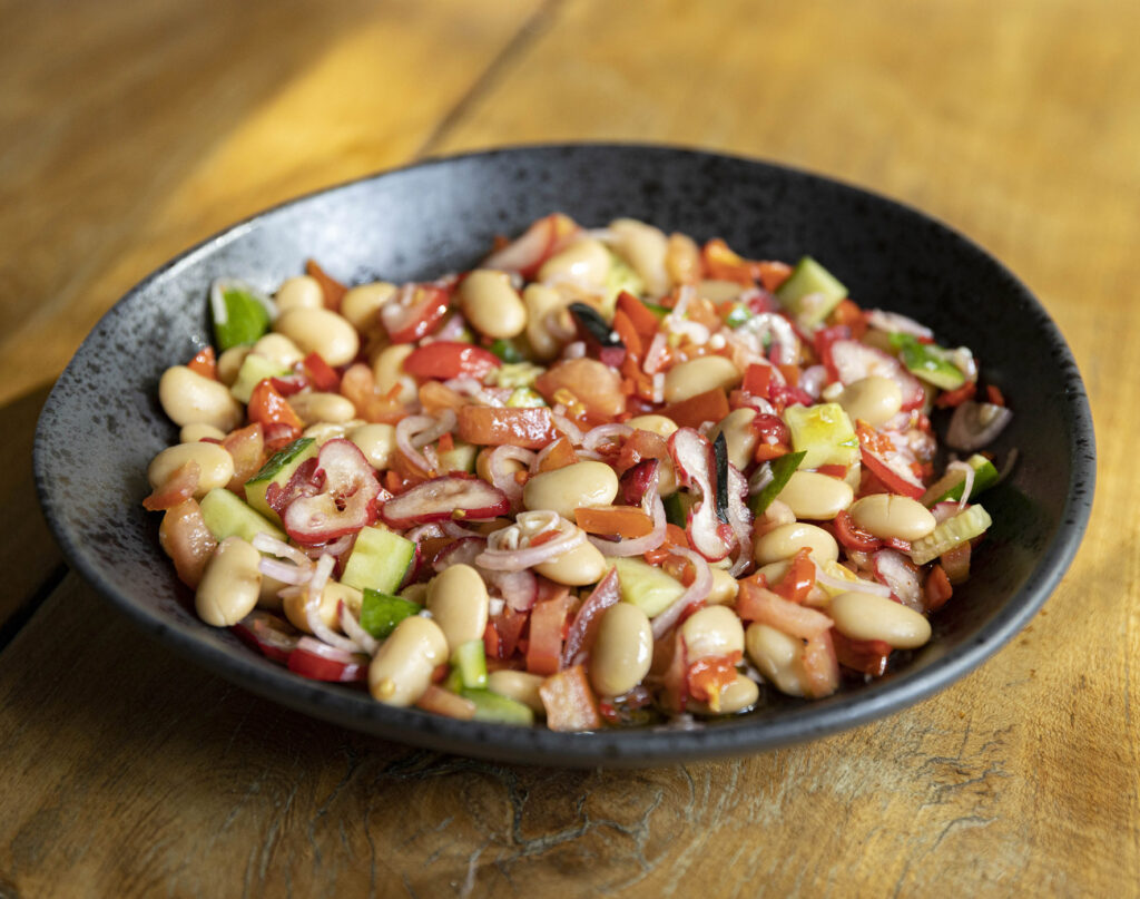A black bowl filled with a white bean salad—white beans, tomatoes, cucumbers, onions, and red peppers—on a wooden table.