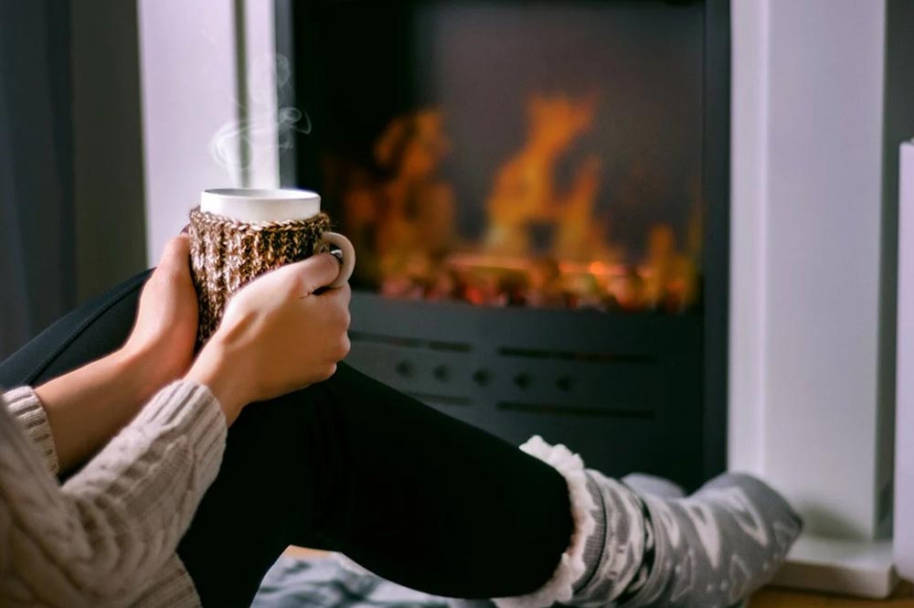 A woman wearing a cosy winter knit and bedsocks hugs a mug in front of a gas fire heater. 