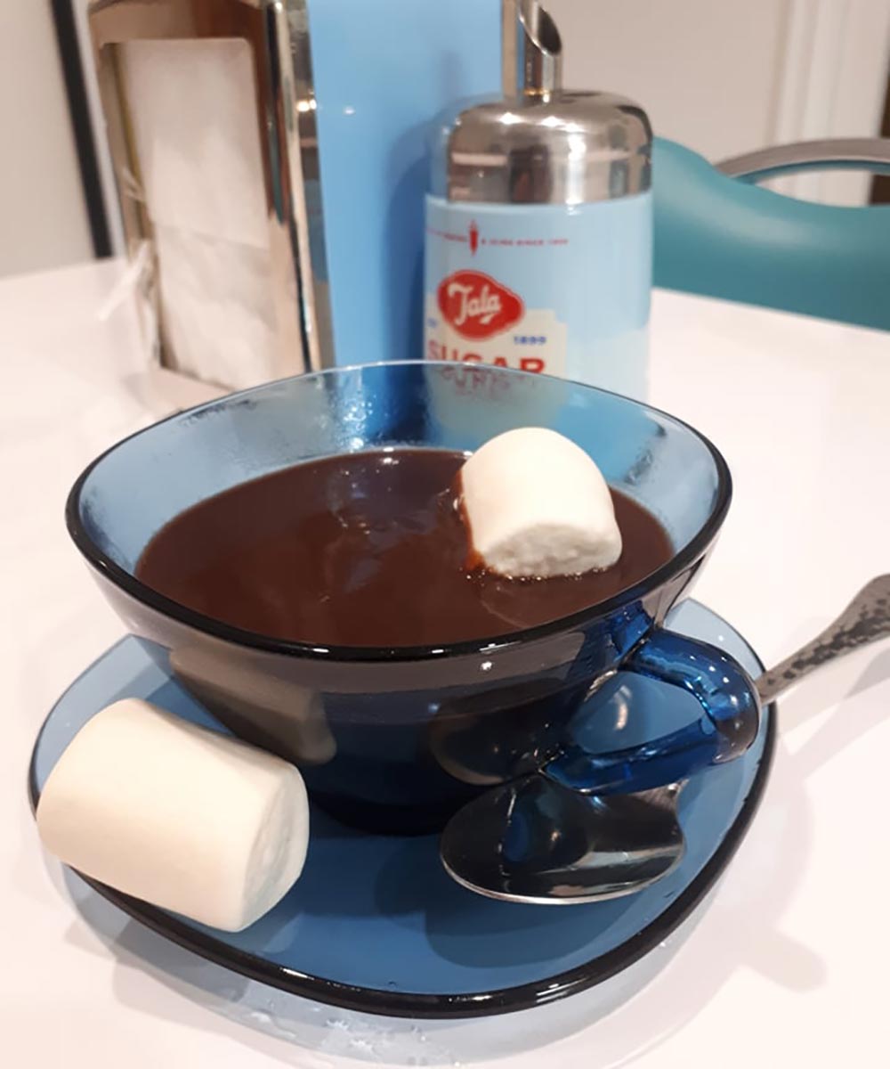 A thick, rich hot chocolate sits in a vintage blue glass, with a marshmallow floating at the top and another to the side. 