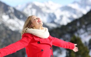 A woman in a red coat and scarf stands out on a snowy day facing the skies with arms wide open and a smile on her face.