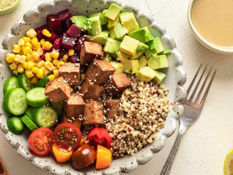 A poke bowl of quinoa, tofu cubes, avocado, sweetcorn, beetroot, cucumber and cherry tomatoes, with a side of dressing and a fork, serving as inspiration for delicious plant-based dishes.