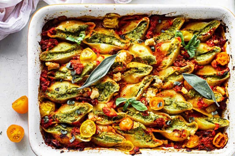 Baked jumbo pasta shells stuffed with spinach and plant-based cheese, featuring a tofu-based filling, topped with tomato sauce, cherry tomatoes, and fresh herbs in a rectangular baking tin.
