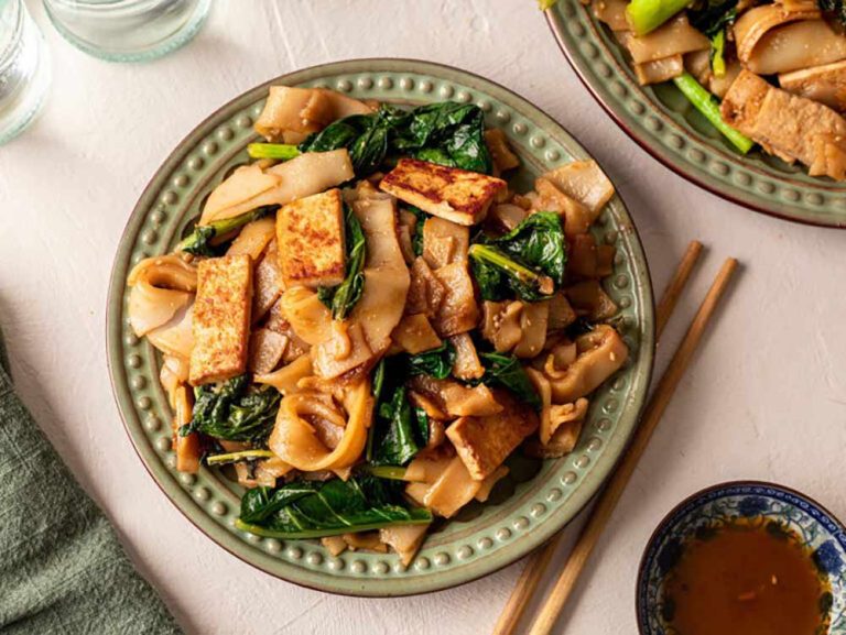 Stir-fried flat rice noodles with tofu and leafy greens on a patterned plate, served with chopsticks and a small bowl of dipping sauce as part of a collection of plant-based meals.