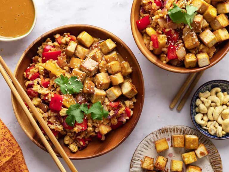 Two bowls of tofu stir-fry with rice, pepper, and cashew nuts, served with chopsticks and extra tofu on the side as a simple plant-based meal.