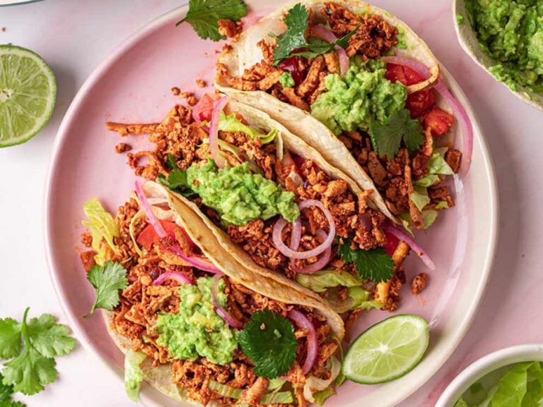 Two plant-based tacos on a pink plate, topped with guacamole, red onion, and coriander, served with lime wedges as part of a vegetarian meal.