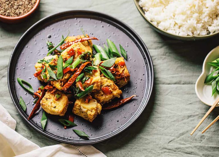 Stir-fried tofu with chilli and shallots on a black plate, served with a bowl of steamed rice as part of our delicious plant-based tofu dishes.