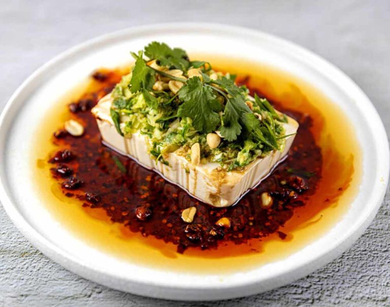 A block of tofu prepared as a plant-based meal, topped with chopped herbs and peanuts in a pool of chilli oil on a white plate.