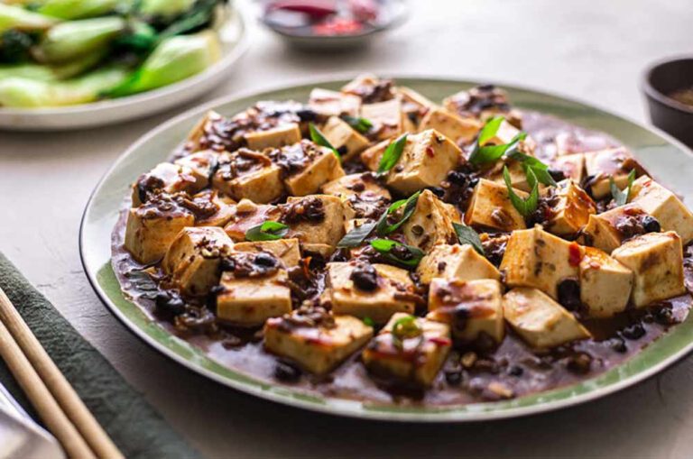 Plate of cubed tofu in a savoury sauce with spring onions, served with a side of pak choi, perfect for plant-based meals.