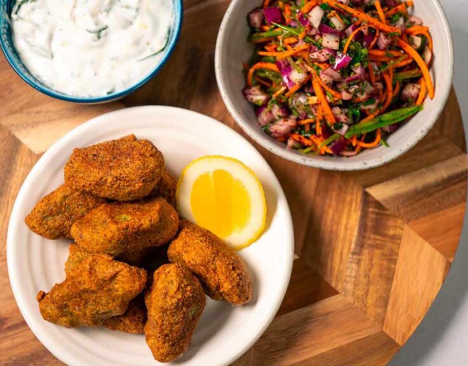 A plate of breaded tofu nuggets with a lemon slice, served with a side of carrot salad and a bowl of dairy-free tzatziki dip.