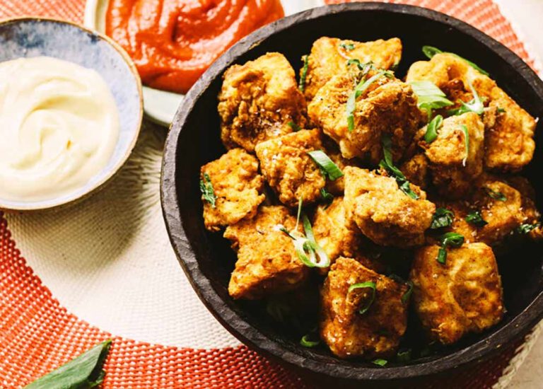 A bowl of crispy fried tofu pieces garnished with sliced shallots, served with side bowls of tomato sauce and vegan mayo, perfect for easy tofu meals.
