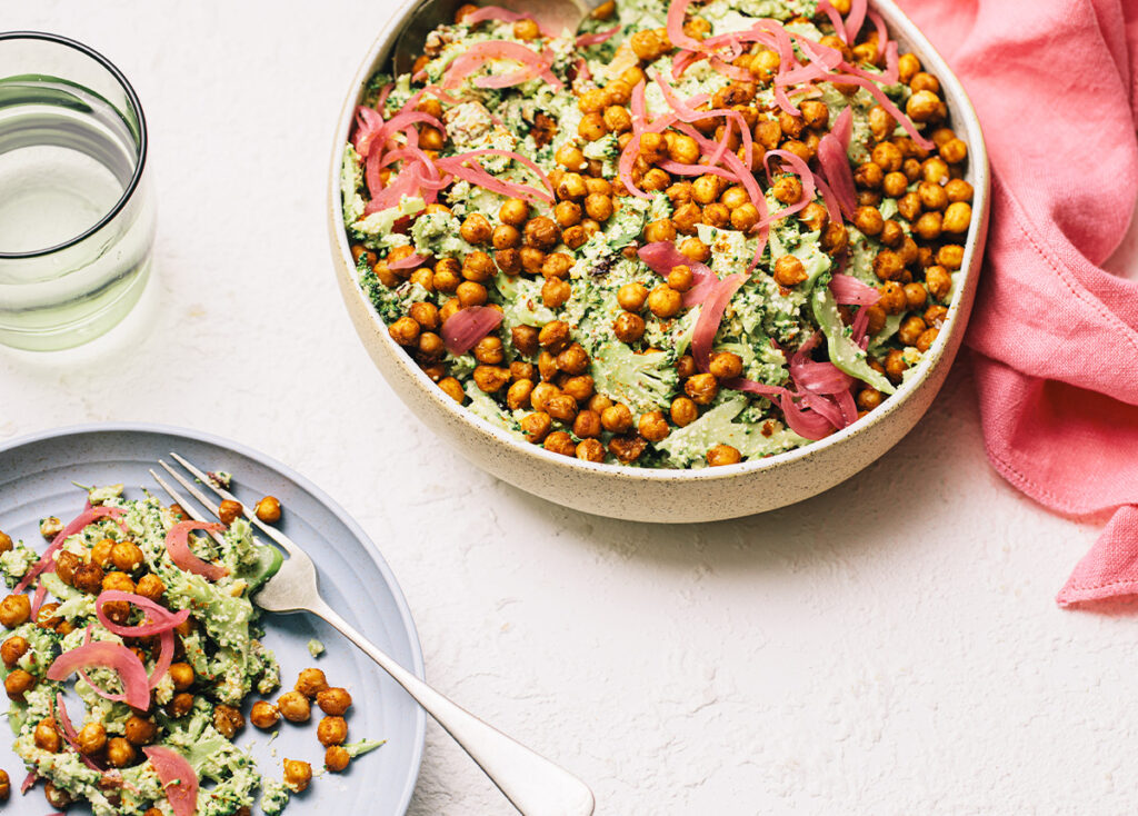 A bowl and plate of salad with crispy chickpeas, plant-based creamy green dressing, pickled onions, broccoli slaw, water, and a pink serviette.