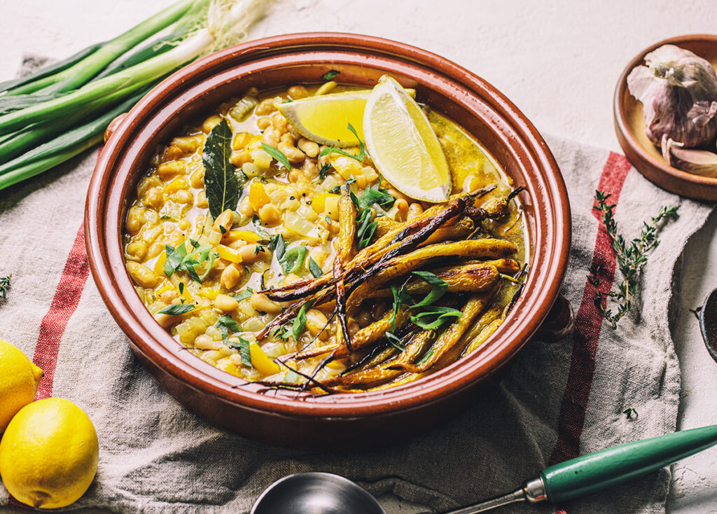 A ceramic bowl of white bean stew topped with roast carrots, lemon wedges, and herbs sits on a striped cloth, with spring onions and garlic beside it.