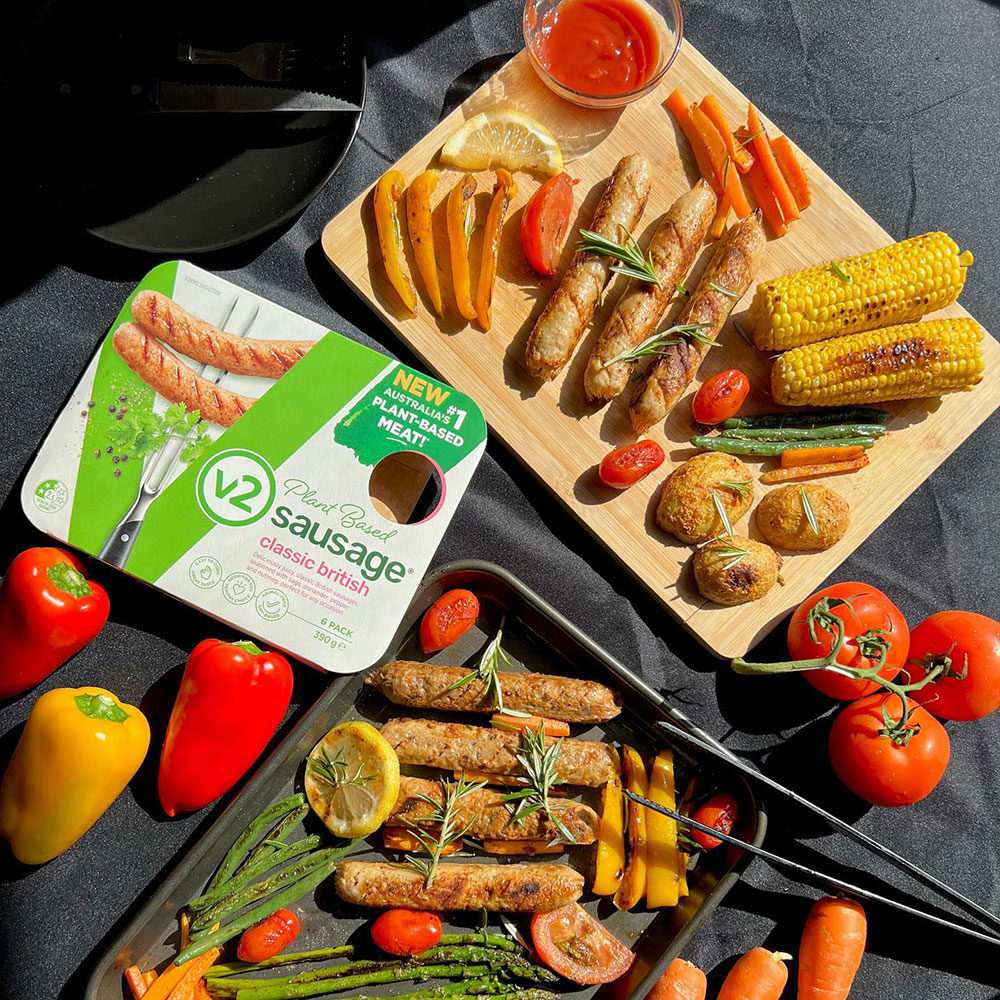 An overhead view of V2 classic British sausages on an oven tray and chopping board, surrounded by a variety of veg for roasting. A V2 pack sits in shot.