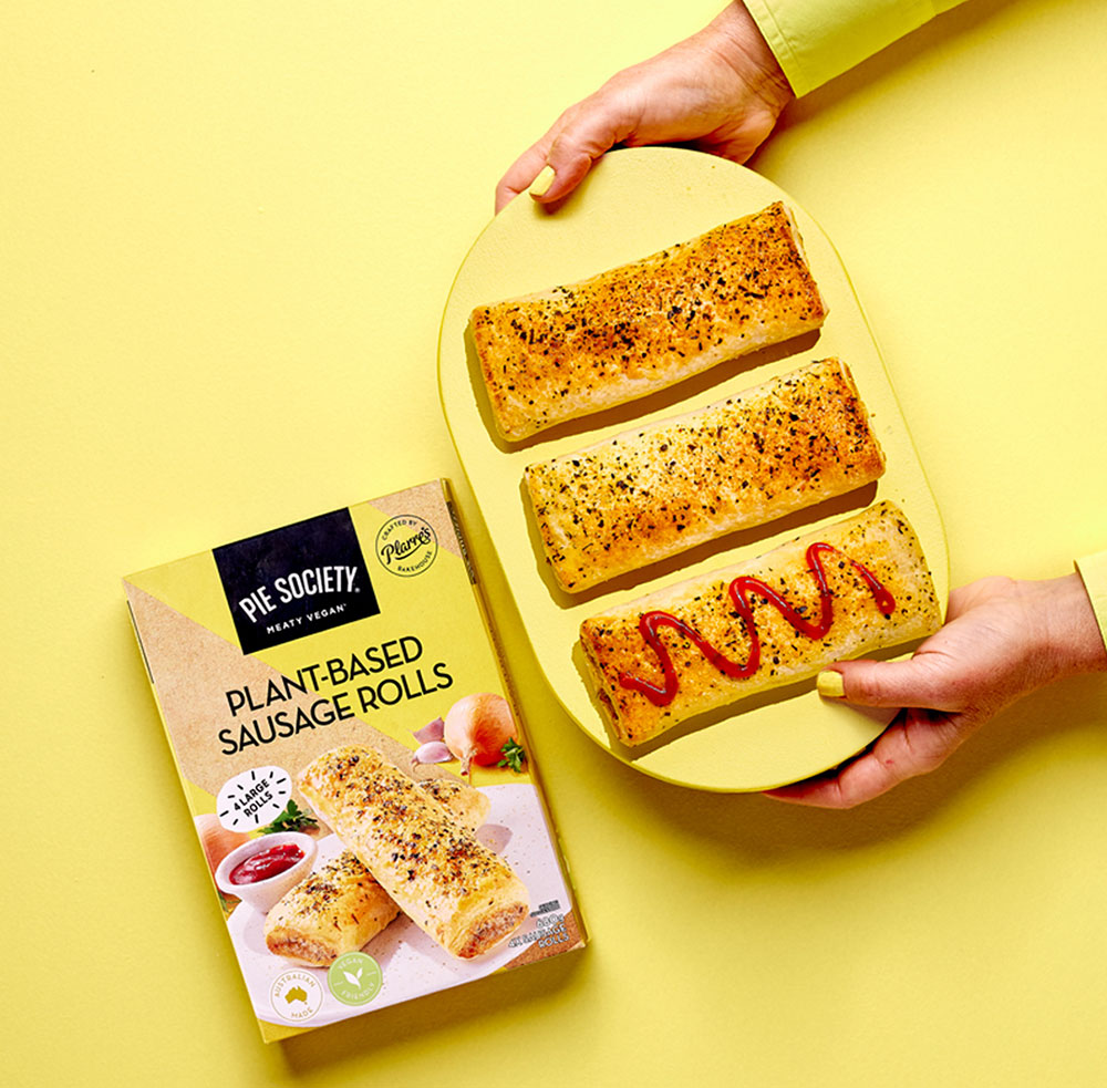 An arm reaches into view holding sausage rolls on a yellow plate. A packet of Pie Society plant-based sausage rolls sits in shot, on a yellow background.