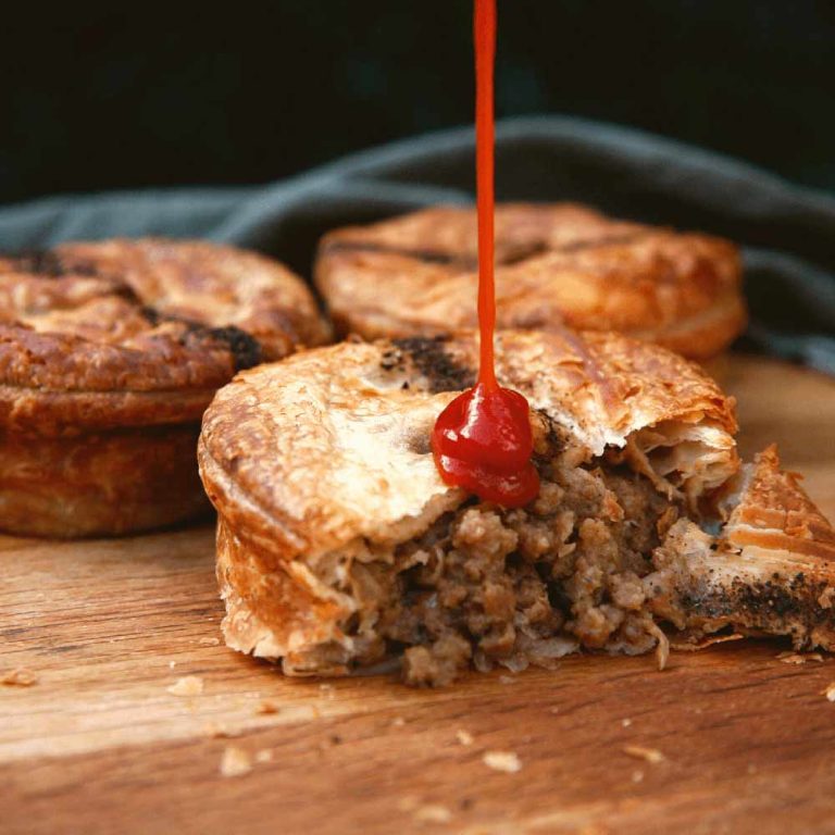 Best vegan sausage rolls & pies: A plant-based pie with a flaky pastry is cut open, revealing its filling. Tomato sauce is being poured onto the pie. Two whole pies, inspired by classic vegan Aussie recipes and packed with tasty plant-based pastry, are in the background on a wooden surface.