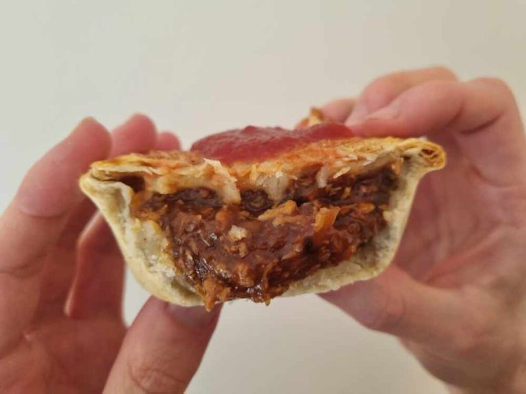 Best vegan sausage rolls & pies: A person holds a half-eaten vegan meat pie topped with tomato sauce, showing the savoury plant-based filling and flaky pastry against a plain background—a tempting treat among the best pastries inspired by Aussie vegan classics.