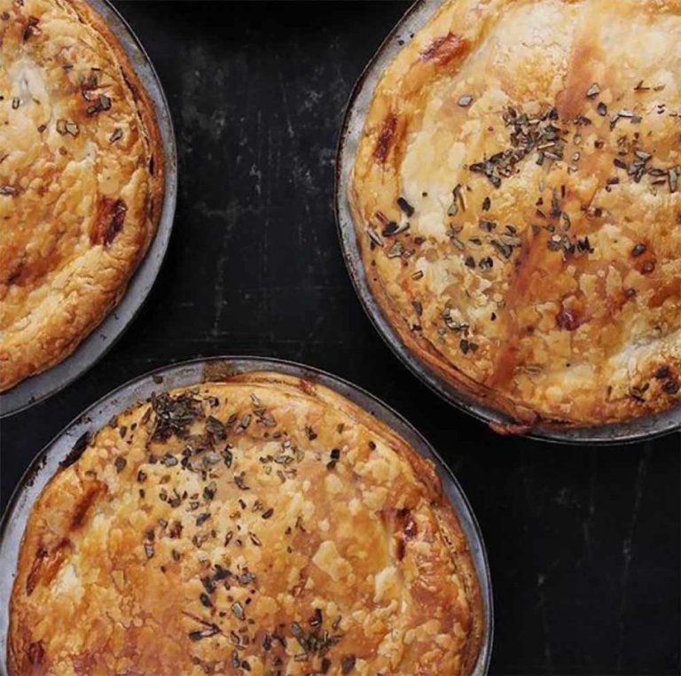 Best vegan sausage rolls & pies: Three round, golden-brown pies with flaky crusts and sprinkled herbs on top, arranged on a dark surface.