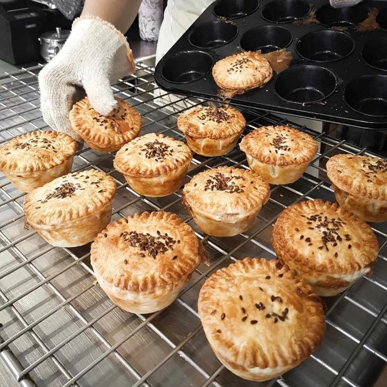 Best vegan sausage rolls & pies: A gloved hand places small baked pies with golden crusts and sprinkled seeds onto a cooling rack beside a muffin tray, highlighting some of the best plant-based pastries in Australia.