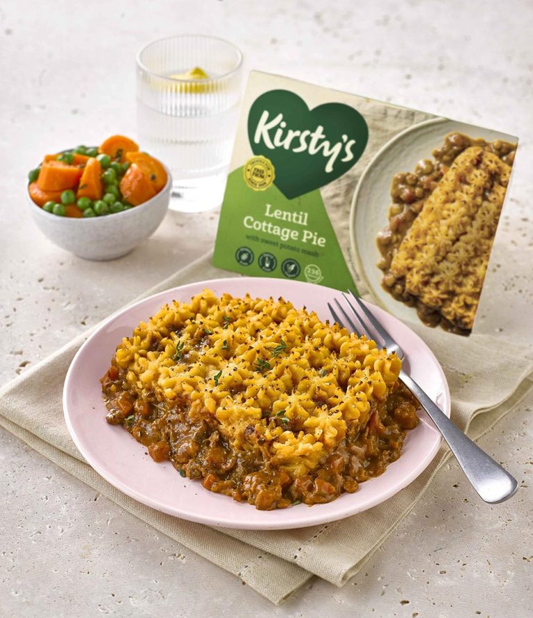 Best vegan sausage rolls & pies: A plate of plant-based lentil shepherd’s pie is served with peas and carrots on the side, a glass of water, and the product packaging labelled Kirstys Lentil Cottage Pie visible in the background.