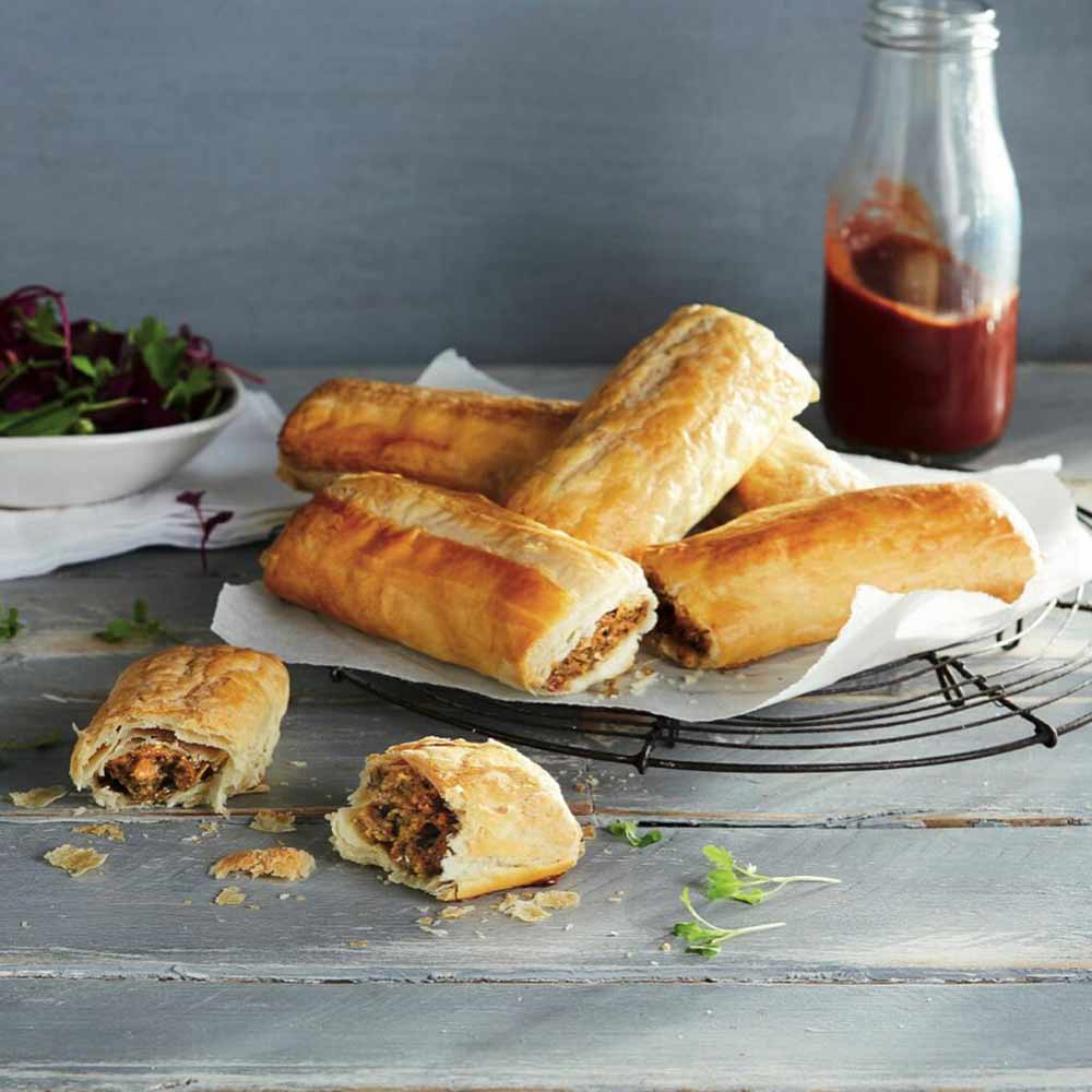 Five top plant-based pastries rest on baking paper, with one partially eaten, all arranged on a cooling rack. A bottle of tomato sauce and a bowl of salad are in the background on a wooden table, making these vegan favourites perfect for any get-together.