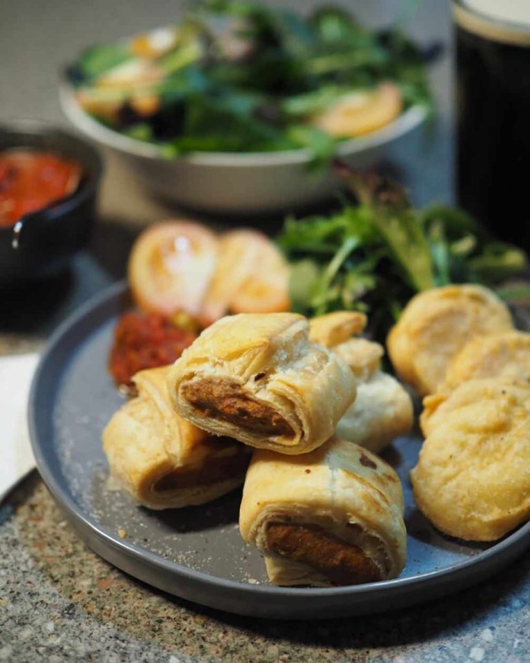 A plate of vegan sausage rolls and vegan pastries with a side of greens, accompanied by a small bowl of sauce; salad and a dark beverage are blurred in the background—ideal for anyone craving top Vegan Aussie classics.