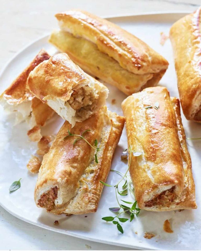 Golden brown vegan sausage rolls with flaky pastry are arranged on a white plate, some broken open to show their plant-based filling, with a few sprigs of fresh herbs as garnish.