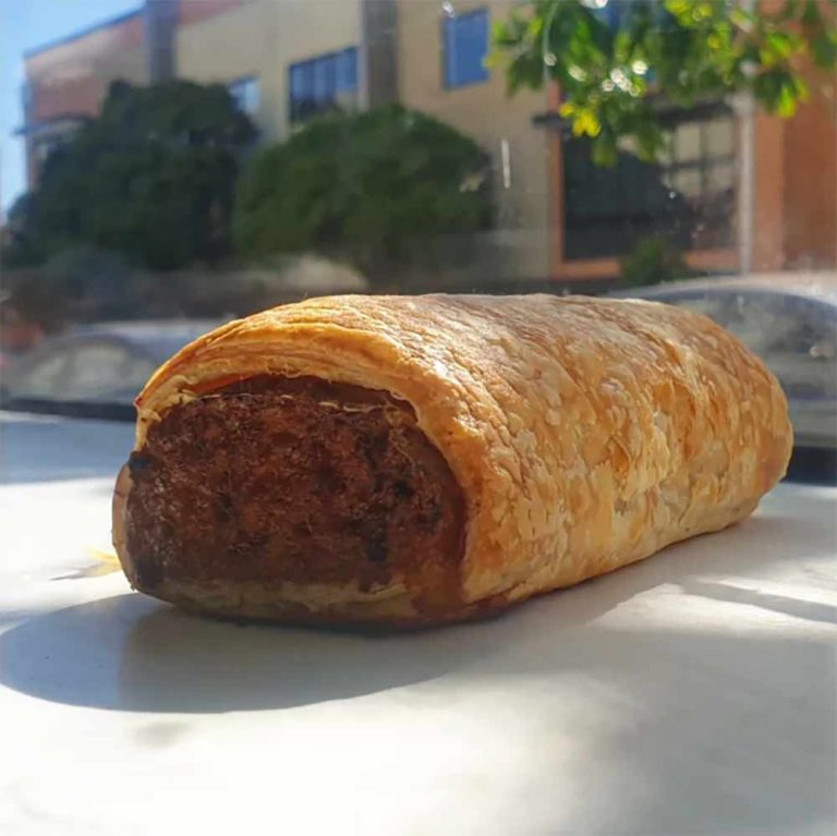A vegan sausage roll with flaky layers of golden pastry rests on a white surface, with blurred buildings and greenery in the background.