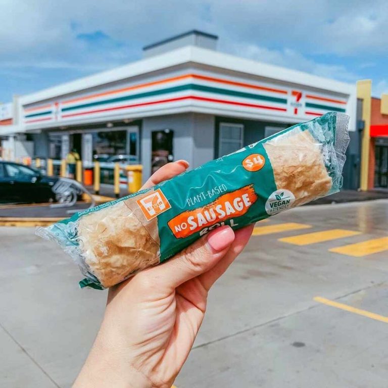 A hand holds a 7-Eleven vegan No Sausage Roll—one of the top vegan Australian favourites—in front of a 7-Eleven shop on a sunny day.