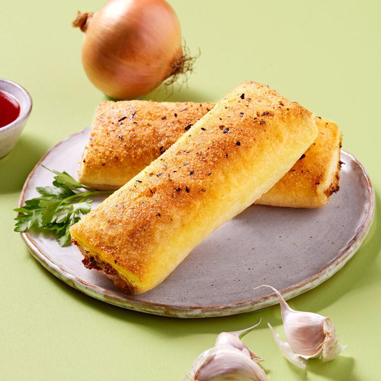 Two golden, seasoned, vegan sausage rolls on a plate with parsley, an onion, garlic cloves, and a small bowl of tomato sauce on a light green surface.