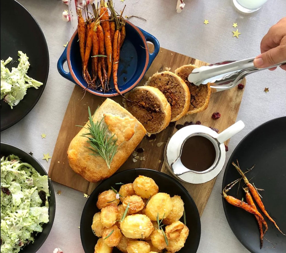 An overhead view of a table with bowls of salad and roast veg, with a pastry-encrusted roast on a wooden serving board in the centre. It has been cut to reveal the centre, and a hand is grabbing one slice with tongs.