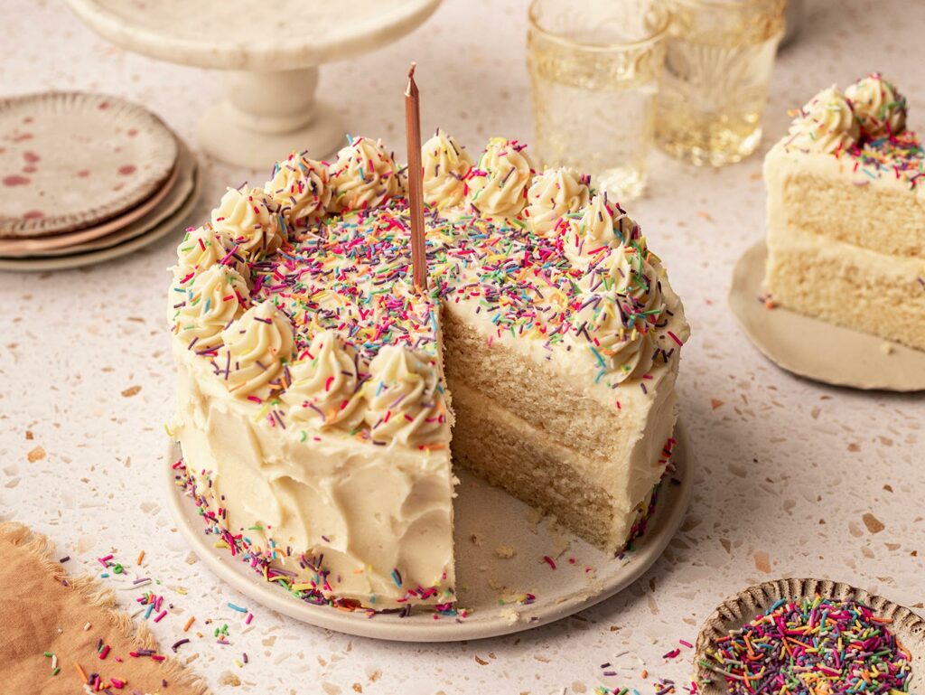 A classic birthday cake: plant-based vanilla sponge cake with vegan white icing, rainbow sprinkles, and a single candle. One slice is removed, with plates, glasses, and a piece of delicious plant-based cake nearby.